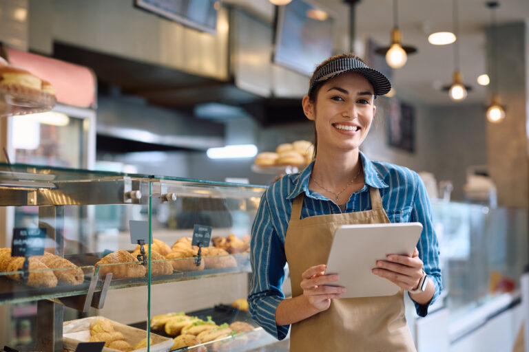Young happy woman small business owner working on digital tablet in a bakery and looking at camera.
